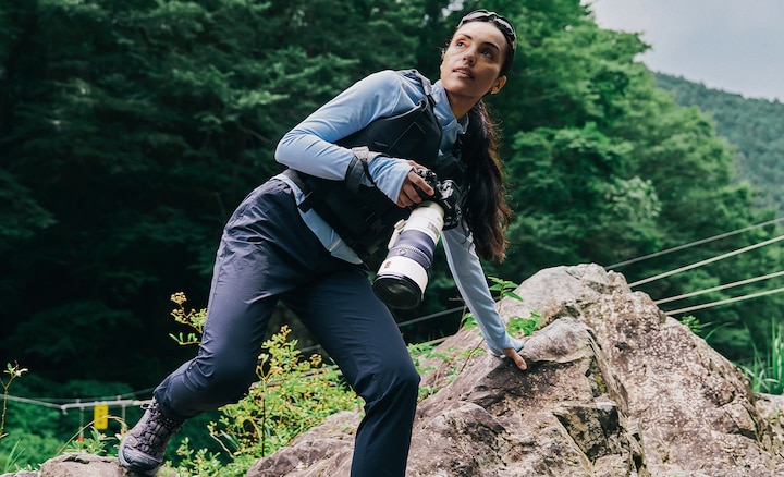 Fotograf, der eine Kamera mit Super-Teleobjektiv trägt und sich einer Hand auf einem Felsen an einem Fluss abstützt